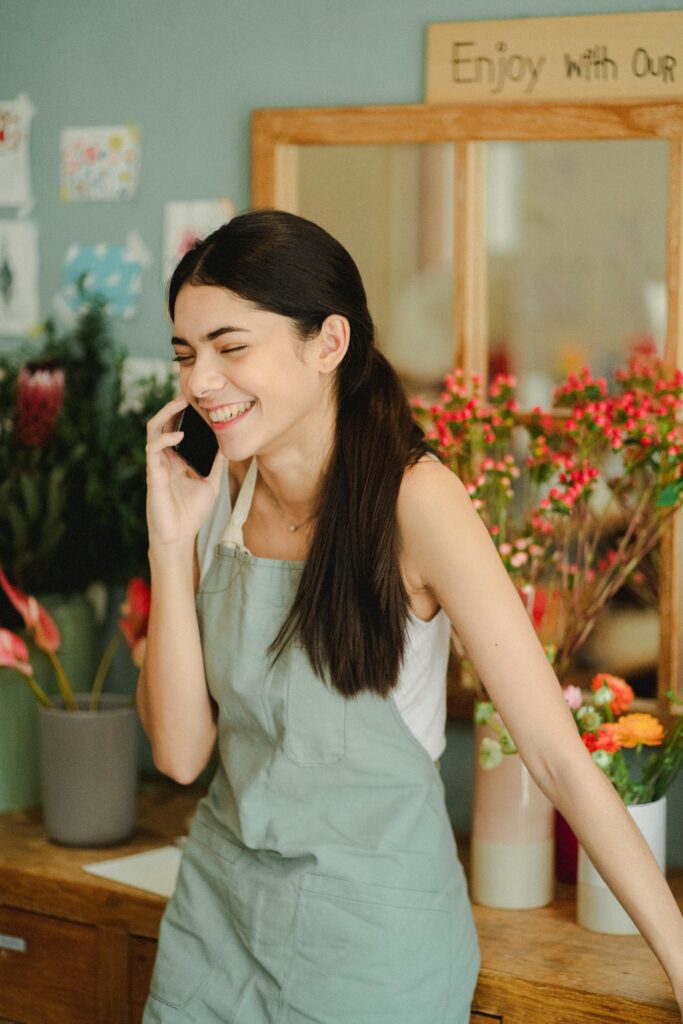 Cheerful female worker with closed eyes wearing apron standing near wooden table with flowers in vase while talking on smartphone in floristry studio